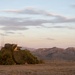 A military vehicle or weapons system is heavily camouflaged with netting and natural vegetation on an open, arid hilltop at dusk or dawn. The equipment appears to include a mounted weapon or optical system along with multiple radio antennas. The scene is set against a backdrop of rolling mountains with a visible moon in the partially cloudy sky.