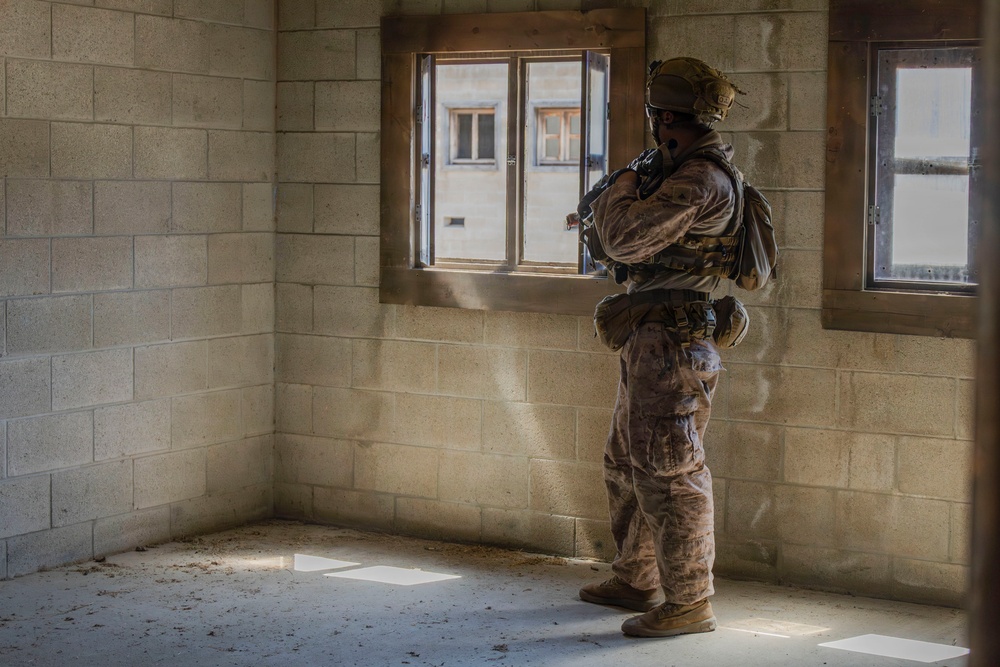 A heavily equipped soldier in full combat gear stands inside a bare concrete block room, positioned beside a window while appearing to observe the exterior. The individual is wearing camouflage uniform, tactical vest, helmet with communications equipment, and carrying a weapon. The setting appears to be a military training facility or urban operations environment.