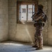 A heavily equipped soldier in full combat gear stands inside a bare concrete block room, positioned beside a window while appearing to observe the exterior. The individual is wearing camouflage uniform, tactical vest, helmet with communications equipment, and carrying a weapon. The setting appears to be a military training facility or urban operations environment.