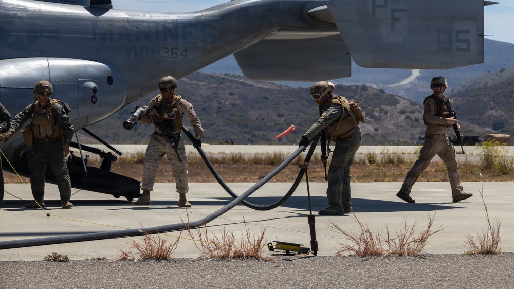 U.S. Marines are conducting a forward arming and refueling point (FARP) operation on a tarmac, handling large fuel hoses connected to a military helicopter. The aircraft visible is marked 'MARINES' and 'VMM-364', identifying it as belonging to Marine Medium Tiltrotor Squadron 364. The setting appears to be a training environment in a dry, hilly terrain consistent with Southern California.