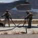 U.S. Marines are conducting a forward arming and refueling point (FARP) operation on a tarmac, handling large fuel hoses connected to a military helicopter. The aircraft visible is marked 'MARINES' and 'VMM-364', identifying it as belonging to Marine Medium Tiltrotor Squadron 364. The setting appears to be a training environment in a dry, hilly terrain consistent with Southern California.