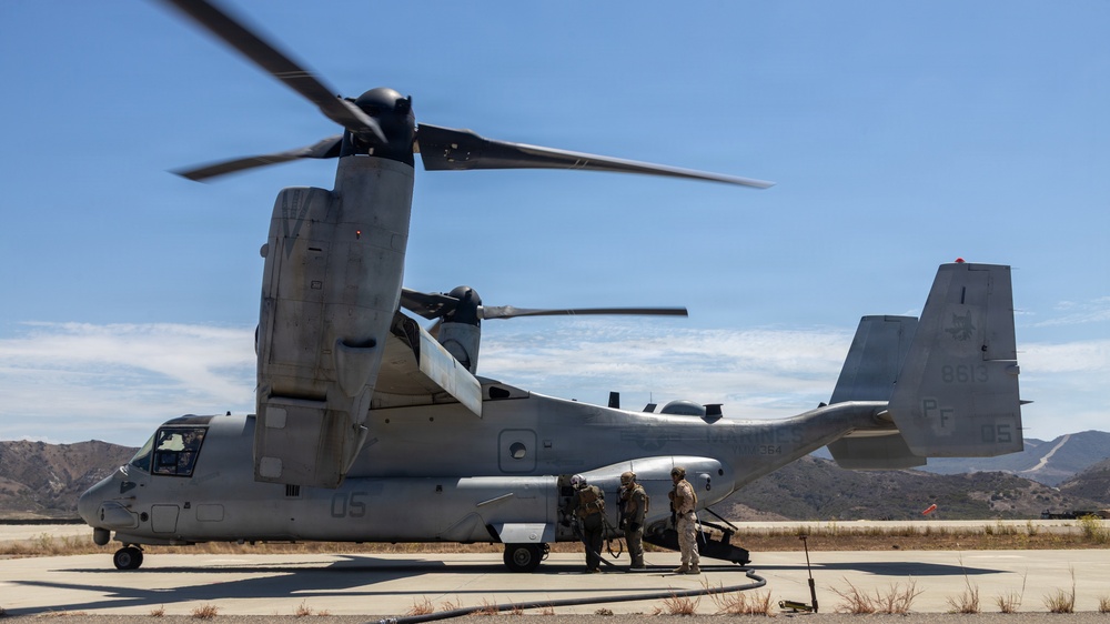 A U.S. Marine Corps MV-22B Osprey tiltrotor aircraft is parked on a tarmac with its rotors spinning, indicating it is either preparing for takeoff or has recently landed. Several military personnel in combat gear are gathered near the rear ramp of the aircraft, likely conducting a pre-mission briefing or boarding procedure. The setting appears to be a desert military installation, consistent with locations such as Marine Corps Base Camp Pendleton or a similar Southern California facility.
