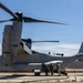 A U.S. Marine Corps MV-22B Osprey tiltrotor aircraft is parked on a tarmac with its rotors spinning, indicating it is either preparing for takeoff or has recently landed. Several military personnel in combat gear are gathered near the rear ramp of the aircraft, likely conducting a pre-mission briefing or boarding procedure. The setting appears to be a desert military installation, consistent with locations such as Marine Corps Base Camp Pendleton or a similar Southern California facility.