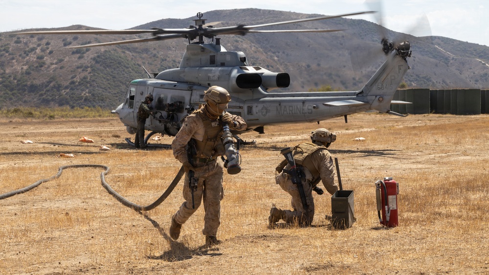 U.S. Marines conduct a forward arming and refueling point (FARP) operation with a UH-1Y Venom helicopter on an arid training field. Personnel in full combat gear are managing fuel hoses and equipment around the idling helicopter, with rotor blades visibly spinning. The setting appears to be a military training installation in a dry, hilly landscape consistent with Southern California.