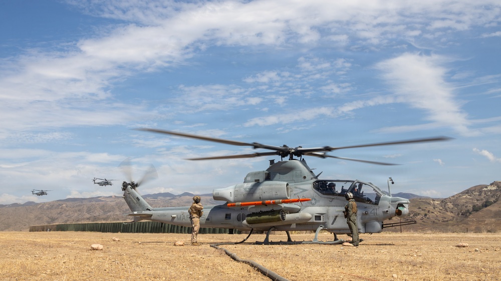 A U.S. Marine Corps AH-1Z Viper attack helicopter sits on an arid forward operating base with rotors spinning, attended by ground crew personnel. The aircraft is armed with visible ordnance including what appear to be AIM-9 Sidewinder missiles and an AGM-114 Hellfire missile. Two additional helicopters are visible in the background taking off or flying nearby, suggesting an active flight operation.