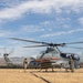 A U.S. Marine Corps AH-1Z Viper attack helicopter sits on an arid forward operating base with rotors spinning, attended by ground crew personnel. The aircraft is armed with visible ordnance including what appear to be AIM-9 Sidewinder missiles and an AGM-114 Hellfire missile. Two additional helicopters are visible in the background taking off or flying nearby, suggesting an active flight operation.
