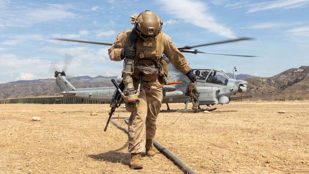 A U.S. Marine in full combat gear walks away from a USMC AH-1Z Viper attack helicopter during what appears to be a forward arming and refueling point (FARP) operation in an arid, hilly training environment. The helicopter's rotors are spinning, indicated by motion blur, and a fuel hose is visible on the ground. The setting appears to be a military training exercise, likely at a base in Southern California based on the terrain.