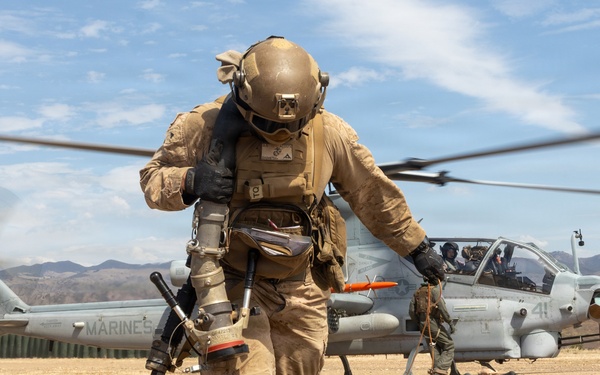 A U.S. Marine in full combat gear walks away from a USMC AH-1Z Viper attack helicopter during what appears to be a forward arming and refueling point (FARP) operation in an arid, hilly training environment. The helicopter's rotors are spinning, indicated by motion blur, and a fuel hose is visible on the ground. The setting appears to be a military training exercise, likely at a base in Southern California based on the terrain.