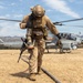 A U.S. Marine in full combat gear walks away from a USMC AH-1Z Viper attack helicopter during what appears to be a forward arming and refueling point (FARP) operation in an arid, hilly training environment. The helicopter's rotors are spinning, indicated by motion blur, and a fuel hose is visible on the ground. The setting appears to be a military training exercise, likely at a base in Southern California based on the terrain.