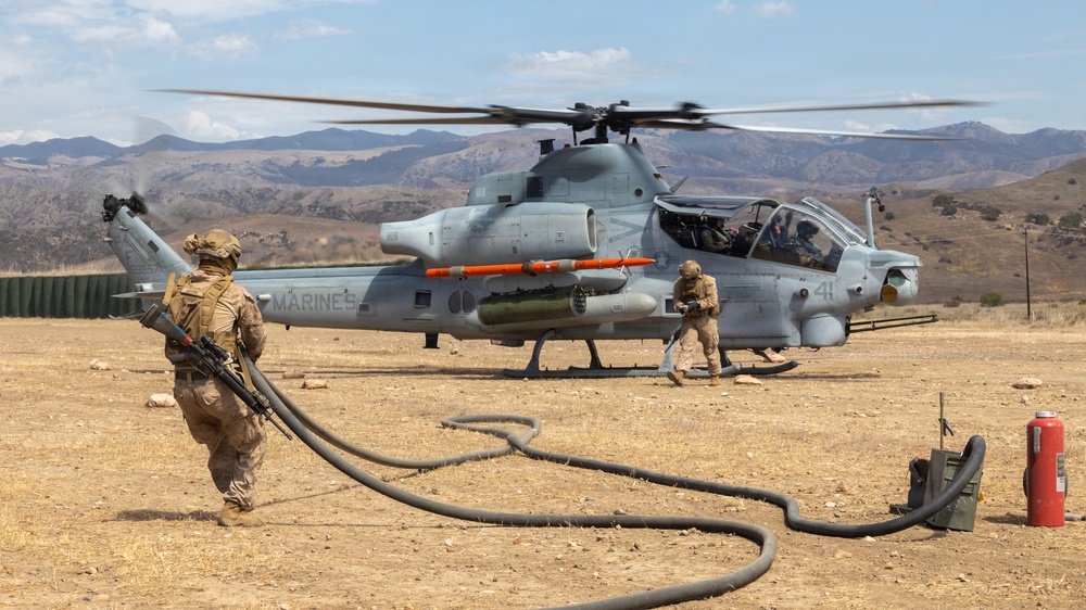 A U.S. Marines AH-1Z Viper attack helicopter is being serviced at a forward arming and refueling point (FARP) in an arid, mountainous terrain. Two Marines in combat gear are conducting ground operations around the aircraft, with one handling a fuel hose and another near the helicopter's landing gear. The helicopter's rotors are spinning, indicating it is in a hot refueling or rearming operation.