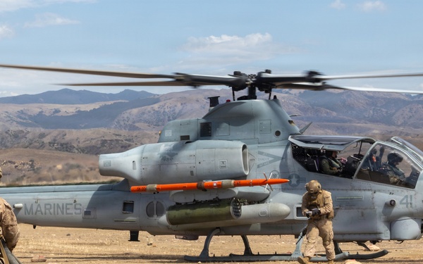 A U.S. Marines AH-1Z Viper attack helicopter is being serviced at a forward arming and refueling point (FARP) in an arid, mountainous terrain. Two Marines in combat gear are conducting ground operations around the aircraft, with one handling a fuel hose and another near the helicopter's landing gear. The helicopter's rotors are spinning, indicating it is in a hot refueling or rearming operation.