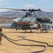 A U.S. Marines AH-1Z Viper attack helicopter is being serviced at a forward arming and refueling point (FARP) in an arid, mountainous terrain. Two Marines in combat gear are conducting ground operations around the aircraft, with one handling a fuel hose and another near the helicopter's landing gear. The helicopter's rotors are spinning, indicating it is in a hot refueling or rearming operation.