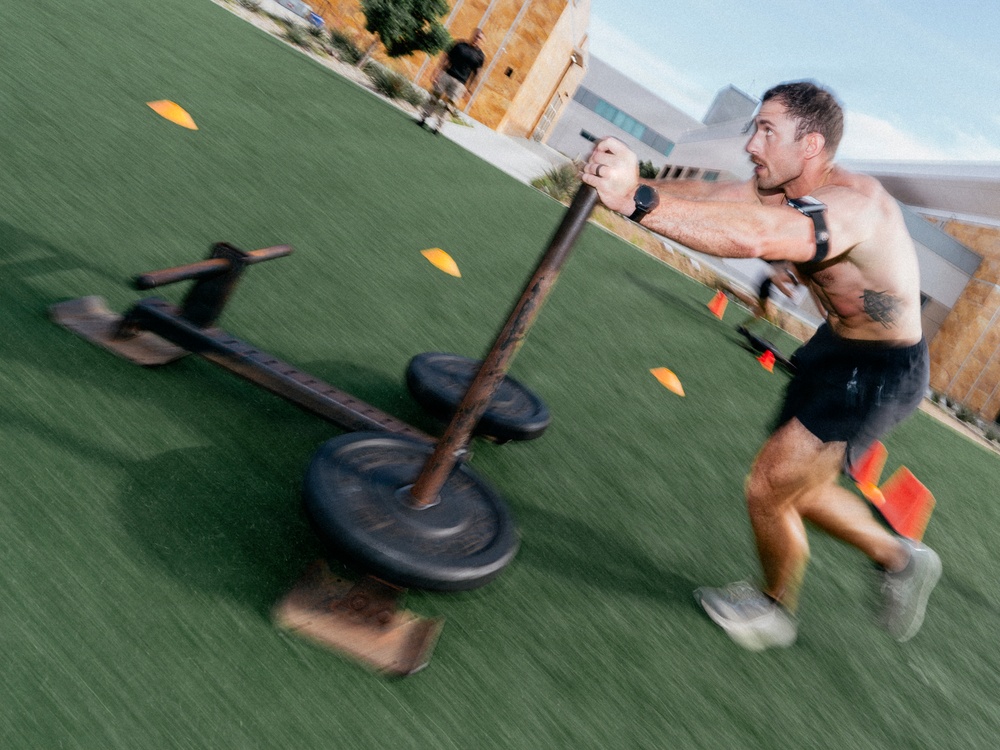 A shirtless athletic man is pushing a weighted sled across an artificial turf field during an outdoor workout session. The image is captured with motion blur, conveying speed and intensity. The setting appears to be an outdoor training facility with marker cones and buildings visible in the background.