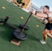 A shirtless athletic man is pushing a weighted sled across an artificial turf field during an outdoor workout session. The image is captured with motion blur, conveying speed and intensity. The setting appears to be an outdoor training facility with marker cones and buildings visible in the background.