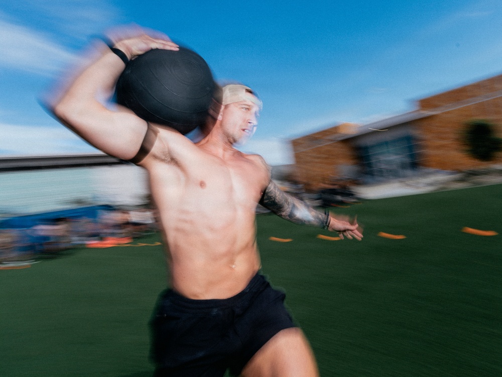 A shirtless, muscular male athlete is captured mid-motion carrying a heavy medicine ball on an outdoor turf field. The image features significant motion blur, conveying speed and intensity during what appears to be a fitness competition or training event. A brick building and blue sky are visible in the blurred background.