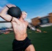 A shirtless, muscular male athlete is captured mid-motion carrying a heavy medicine ball on an outdoor turf field. The image features significant motion blur, conveying speed and intensity during what appears to be a fitness competition or training event. A brick building and blue sky are visible in the blurred background.