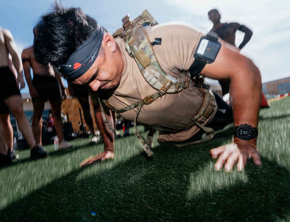 A man is performing push-ups on artificial turf while wearing a tactical/military-style weighted vest or ruck pack over a tan t-shirt. He is also wearing a black headband and a digital sports watch on his right wrist. The image appears to be taken at an outdoor fitness event or military-style competition with other participants visible in the background.
