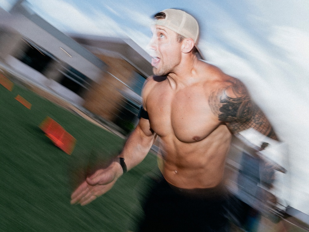 A shirtless, athletic male is captured mid-sprint on what appears to be an outdoor turf field. The image uses motion blur to convey speed and intensity, with the subject wearing a backwards baseball cap and displaying extensive tattoos on his right arm and shoulder. His expression is intense and open-mouthed, suggesting maximum physical exertion.