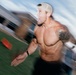 A shirtless, athletic male is captured mid-sprint on what appears to be an outdoor turf field. The image uses motion blur to convey speed and intensity, with the subject wearing a backwards baseball cap and displaying extensive tattoos on his right arm and shoulder. His expression is intense and open-mouthed, suggesting maximum physical exertion.