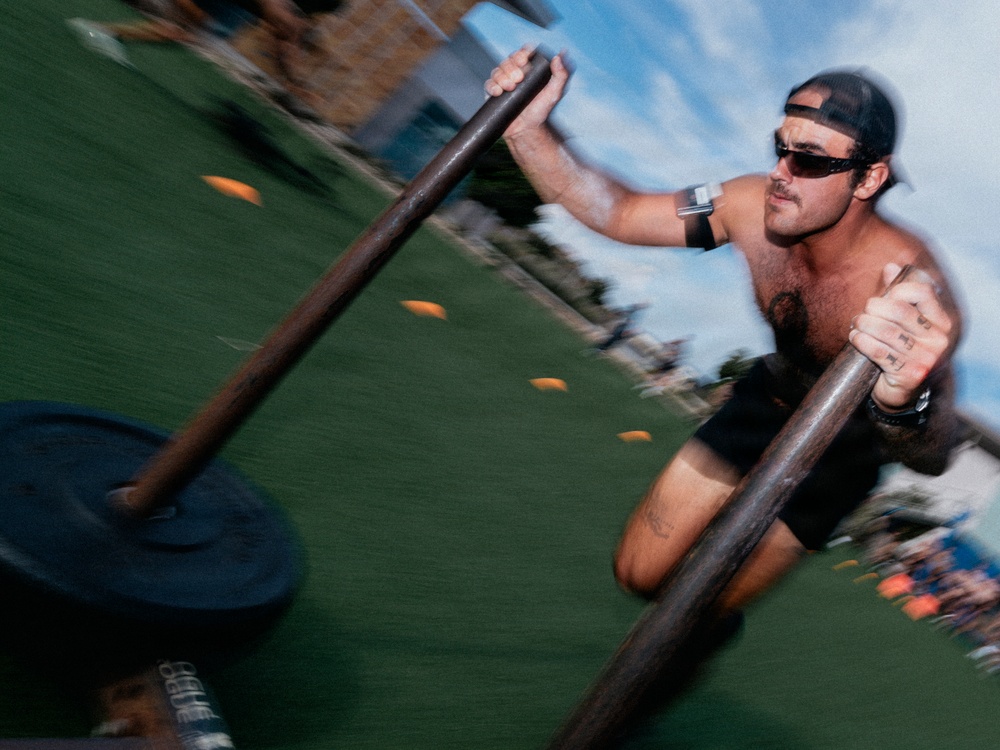 A shirtless man wearing a backwards baseball cap and sunglasses is pushing a weighted sled across an artificial turf field. The image is captured with motion blur, conveying speed and intensity during what appears to be an outdoor fitness or athletic training session. Orange cones are visible in the background, suggesting a structured training course.