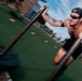 A shirtless man wearing a backwards baseball cap and sunglasses is pushing a weighted sled across an artificial turf field. The image is captured with motion blur, conveying speed and intensity during what appears to be an outdoor fitness or athletic training session. Orange cones are visible in the background, suggesting a structured training course.