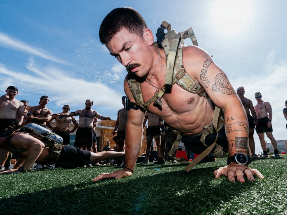 A shirtless muscular man wearing a tactical/military-style chest rig is performing push-ups on an artificial turf field. He has visible tattoos on his arms and shoulders, and wears a watch on his left wrist. A group of similarly shirtless individuals can be seen in the background, suggesting a group fitness or military-style training event.