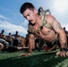 A shirtless muscular man wearing a tactical/military-style chest rig is performing push-ups on an artificial turf field. He has visible tattoos on his arms and shoulders, and wears a watch on his left wrist. A group of similarly shirtless individuals can be seen in the background, suggesting a group fitness or military-style training event.
