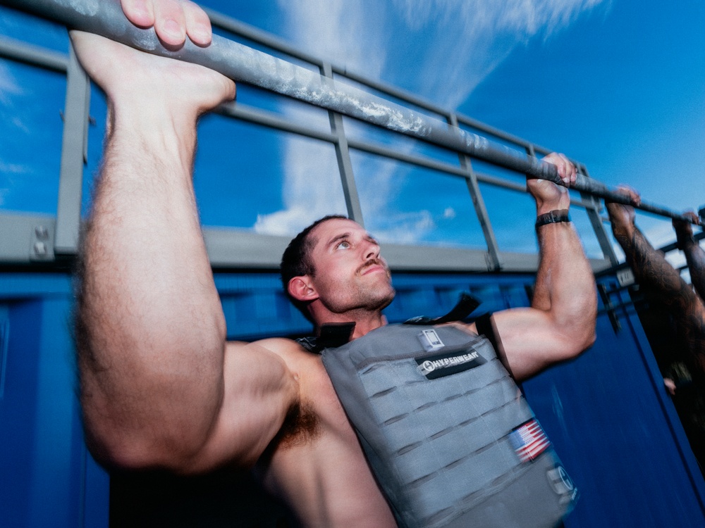 A muscular man is performing pull-ups on a metal bar outdoors while wearing a weighted vest. He is shirtless beneath the vest and has his legs raised, suggesting an advanced exercise technique. The photo is taken from a low angle against a bright blue sky.