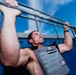 A muscular man is performing pull-ups on a metal bar outdoors while wearing a weighted vest. He is shirtless beneath the vest and has his legs raised, suggesting an advanced exercise technique. The photo is taken from a low angle against a bright blue sky.