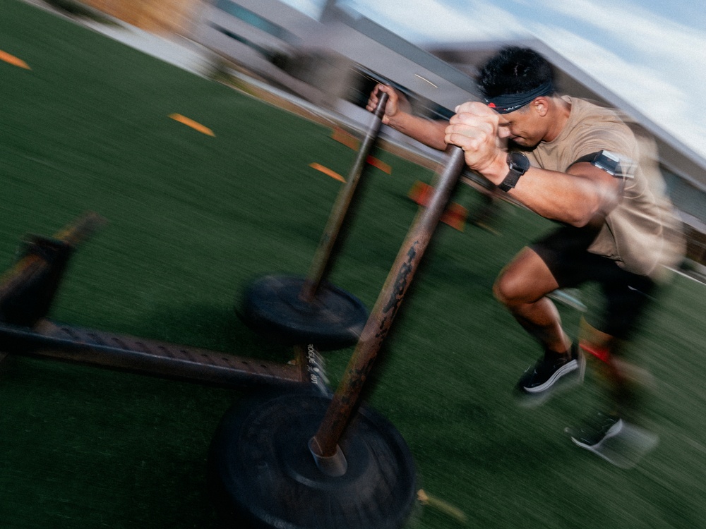 A muscular athlete is pushing a weighted sled across an artificial turf field during an intense training session. The motion blur effect captures the speed and effort of the exercise, emphasizing the physical exertion involved. The setting appears to be an outdoor athletic training facility or sports field.