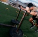 A muscular athlete is pushing a weighted sled across an artificial turf field during an intense training session. The motion blur effect captures the speed and effort of the exercise, emphasizing the physical exertion involved. The setting appears to be an outdoor athletic training facility or sports field.