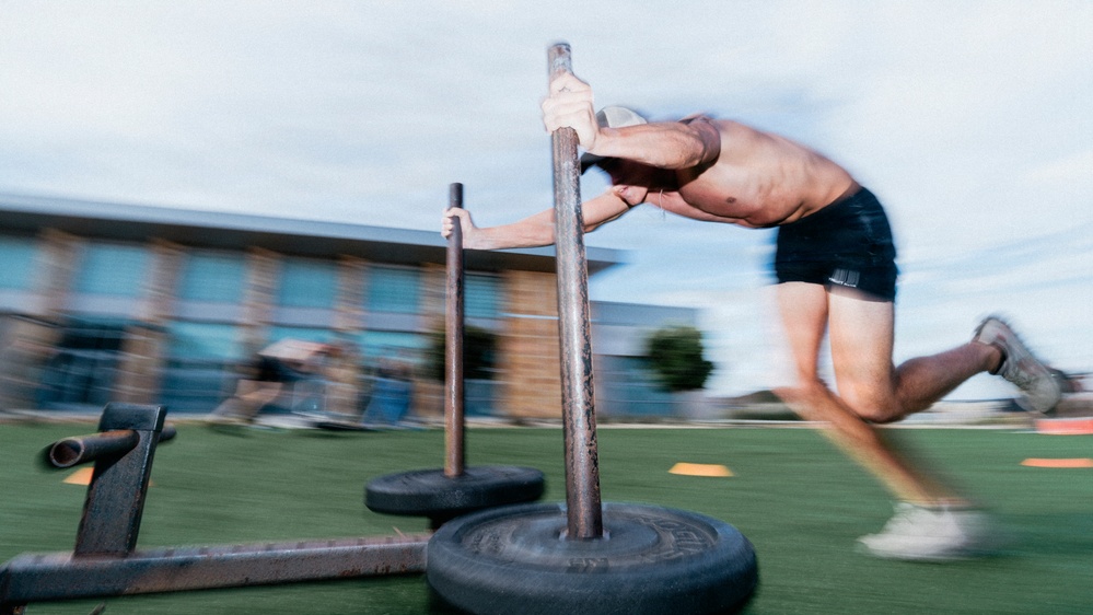 A shirtless male athlete is pushing a weighted sled across an outdoor turf training area. The image is captured with motion blur, conveying speed and intensity during what appears to be a functional fitness or athletic conditioning workout. The setting appears to be an outdoor training facility with a building visible in the background.