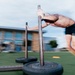 A shirtless male athlete is pushing a weighted sled across an outdoor turf training area. The image is captured with motion blur, conveying speed and intensity during what appears to be a functional fitness or athletic conditioning workout. The setting appears to be an outdoor training facility with a building visible in the background.
