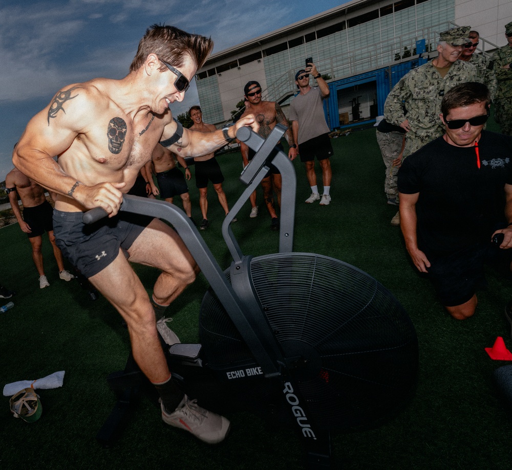 A shirtless, heavily tattooed man is intensely using a Rogue Echo Bike on an outdoor turf area, surrounded by a group of onlookers. Several individuals in Navy digital camouflage uniforms are visible observing the activity, along with other shirtless participants. The setting appears to be a military fitness event or demonstration.