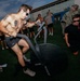 A shirtless, heavily tattooed man is intensely using a Rogue Echo Bike on an outdoor turf area, surrounded by a group of onlookers. Several individuals in Navy digital camouflage uniforms are visible observing the activity, along with other shirtless participants. The setting appears to be a military fitness event or demonstration.