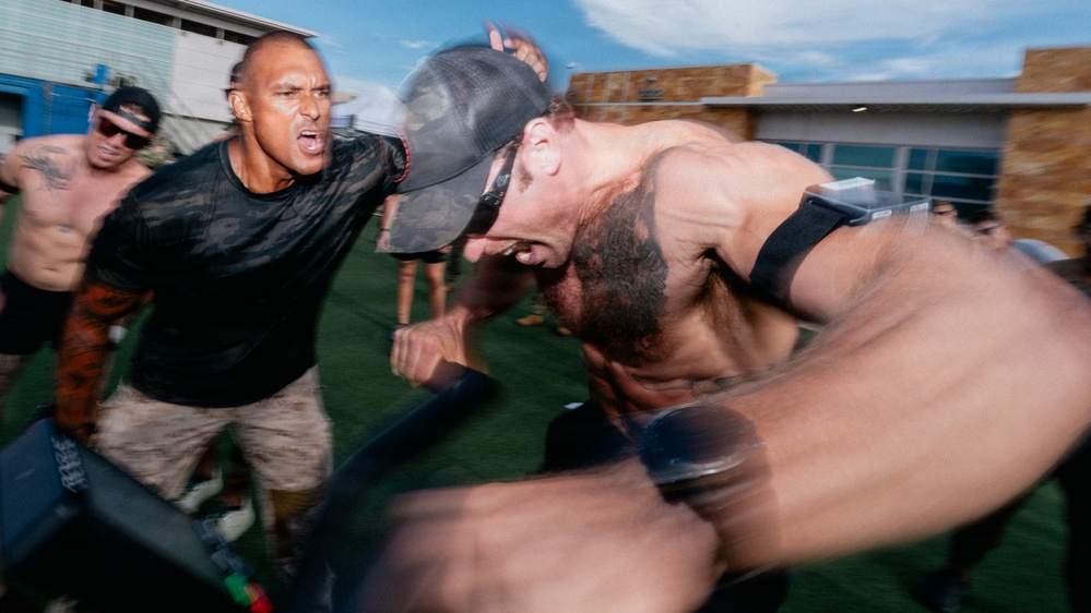 A high-intensity outdoor fitness or training event captured with motion blur, showing multiple participants pushing through a physically demanding exercise. The foreground subject, shirtless and wearing a baseball cap, appears to be pushing or pulling heavy equipment with extreme exertion. A coach or trainer in a black shirt is shouting encouragement or instruction nearby.