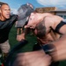 A high-intensity outdoor fitness or training event captured with motion blur, showing multiple participants pushing through a physically demanding exercise. The foreground subject, shirtless and wearing a baseball cap, appears to be pushing or pulling heavy equipment with extreme exertion. A coach or trainer in a black shirt is shouting encouragement or instruction nearby.