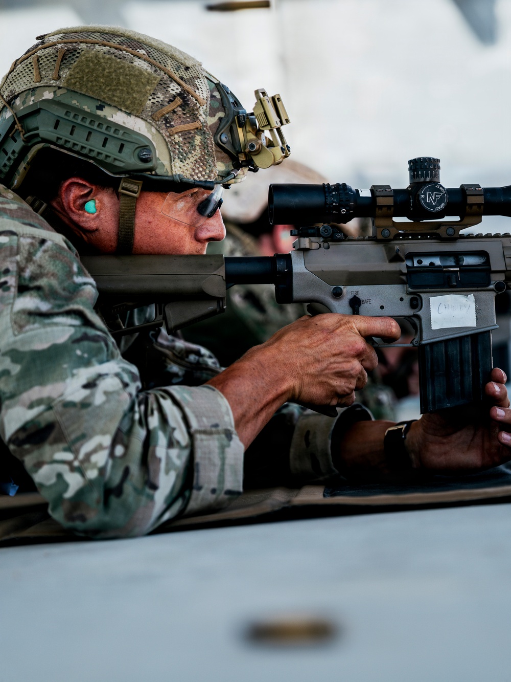 A military service member in full combat gear is aiming a rifle equipped with a high-powered scope over a surface, likely a vehicle. The individual is wearing a multicam camouflage uniform, tactical helmet with night vision mount, ear protection, and ballistic glasses. The image appears to depict a training or operational scenario.