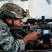 A military service member in full combat gear is aiming a rifle equipped with a high-powered scope over a surface, likely a vehicle. The individual is wearing a multicam camouflage uniform, tactical helmet with night vision mount, ear protection, and ballistic glasses. The image appears to depict a training or operational scenario.