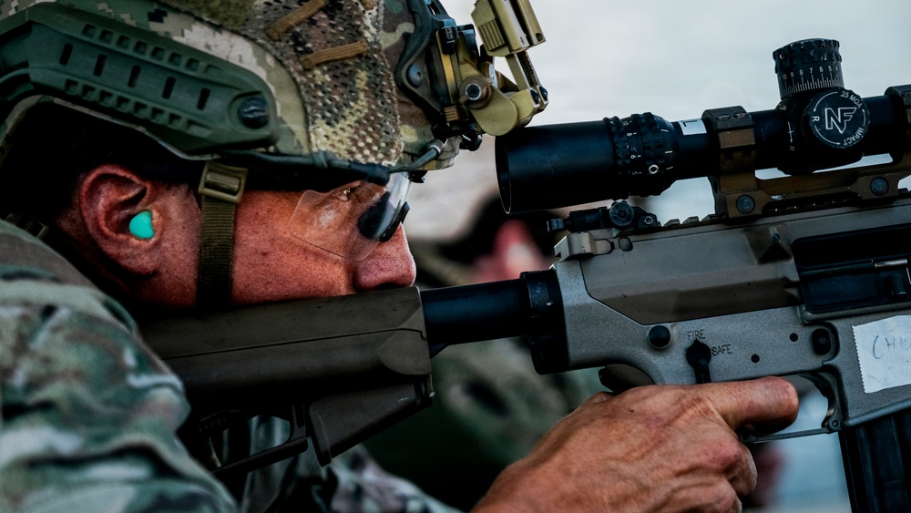 A soldier in modern military camouflage (MultiCam/OCP pattern) is seen aiming down the scope of a rifle during what appears to be a training exercise or range qualification. The shooter is equipped with advanced optics, a tactical helmet with mounted devices, and hearing protection. The close-up composition emphasizes the precision and focus of the marksman.