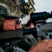 A soldier in modern military camouflage (MultiCam/OCP pattern) is seen aiming down the scope of a rifle during what appears to be a training exercise or range qualification. The shooter is equipped with advanced optics, a tactical helmet with mounted devices, and hearing protection. The close-up composition emphasizes the precision and focus of the marksman.