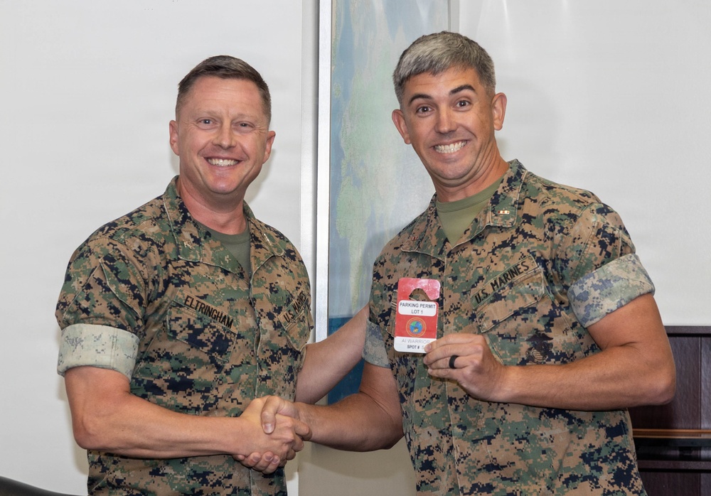 Two U.S. Marines in MARPAT camouflage uniforms are shaking hands while one presents a red parking permit placard to the other. The Marine on the left is identified by his name tape as 'ELTRINGHAM.' Both are smiling, suggesting a ceremonial or recognition moment.