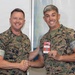Two U.S. Marines in MARPAT camouflage uniforms are shaking hands while one presents a red parking permit placard to the other. The Marine on the left is identified by his name tape as 'ELTRINGHAM.' Both are smiling, suggesting a ceremonial or recognition moment.