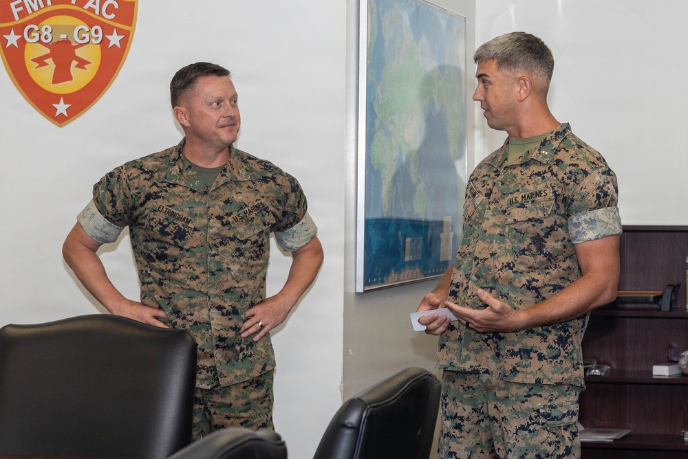 Two U.S. Marines in MARPAT camouflage uniforms are engaged in conversation inside what appears to be a military office. The setting includes a FMFPAC G8-G9 unit insignia on the wall and a world map in the background. The Marine on the right appears to hold a higher rank based on visible collar insignia.