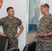Two U.S. Marines in MARPAT camouflage uniforms are engaged in conversation inside what appears to be a military office. The setting includes a FMFPAC G8-G9 unit insignia on the wall and a world map in the background. The Marine on the right appears to hold a higher rank based on visible collar insignia.