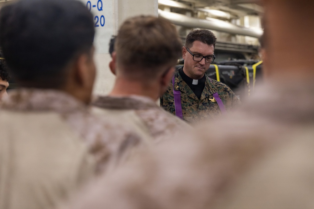 A military chaplain wearing MARPAT camouflage uniform with a purple stole and clerical collar is conducting what appears to be a religious service or prayer with a group of Marines. The setting appears to be inside a naval vessel, likely a well deck or vehicle storage area, indicated by the industrial surroundings and military equipment visible in the background. The chaplain is looking downward, suggesting a moment of prayer or reflection.