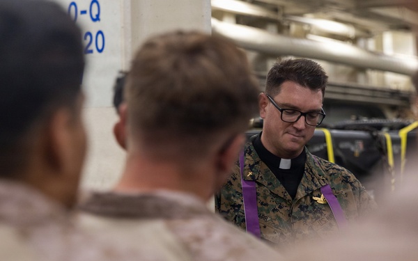 A military chaplain wearing MARPAT camouflage uniform with a purple stole and clerical collar is conducting what appears to be a religious service or prayer with a group of Marines. The setting appears to be inside a naval vessel, likely a well deck or vehicle storage area, indicated by the industrial surroundings and military equipment visible in the background. The chaplain is looking downward, suggesting a moment of prayer or reflection.