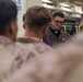 A military chaplain wearing MARPAT camouflage uniform with a purple stole and clerical collar is conducting what appears to be a religious service or prayer with a group of Marines. The setting appears to be inside a naval vessel, likely a well deck or vehicle storage area, indicated by the industrial surroundings and military equipment visible in the background. The chaplain is looking downward, suggesting a moment of prayer or reflection.