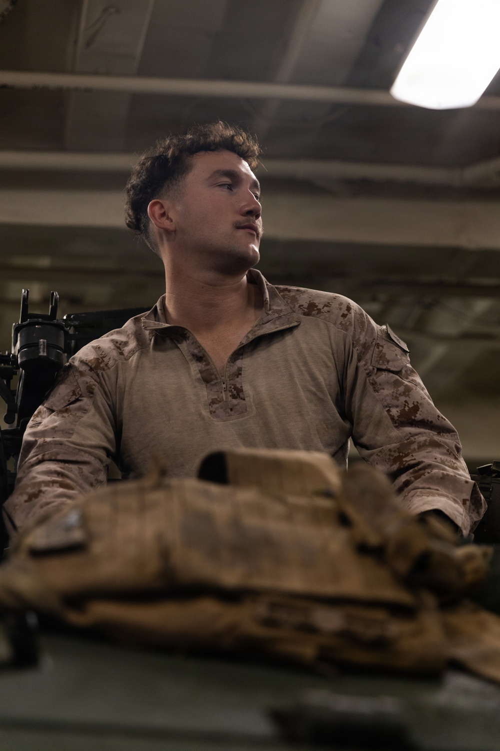 A young male individual wearing USMC MARPAT desert camouflage combat shirt is seated in what appears to be an indoor military or industrial environment. Tactical gear and equipment are visible in the foreground, and a weapon system is partially visible to the left. The subject is looking off to one side under artificial overhead lighting.