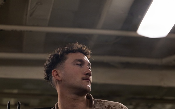 A young male individual wearing USMC MARPAT desert camouflage combat shirt is seated in what appears to be an indoor military or industrial environment. Tactical gear and equipment are visible in the foreground, and a weapon system is partially visible to the left. The subject is looking off to one side under artificial overhead lighting.